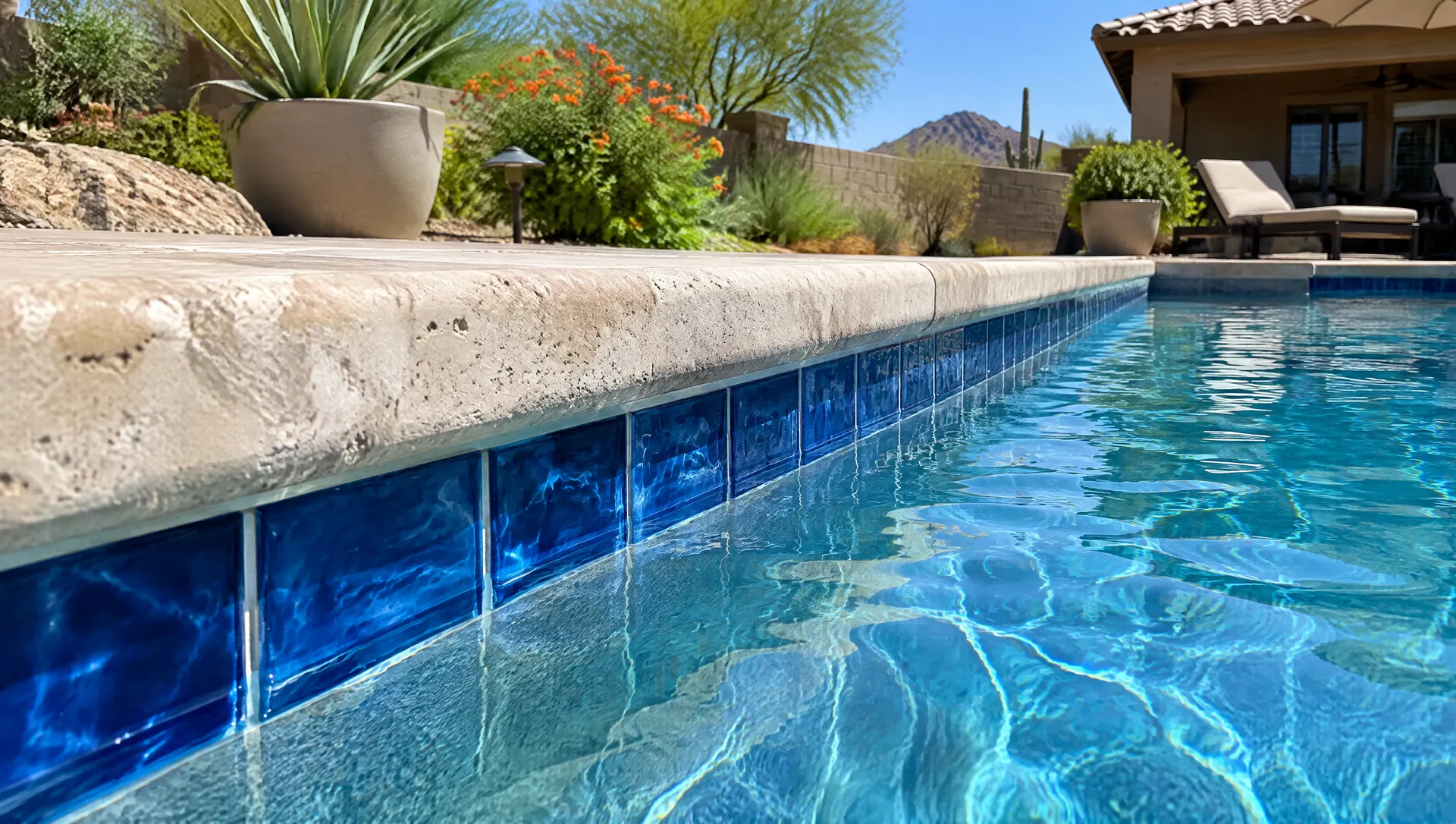 Close view along a Phoenix backyard pool edge with new glossy deep blue glass waterline tile set just under tan travertine coping, clear water lapping the tile, no calcium stains